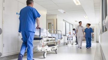Healthcare workers in a hospital corridor, with a person pushing a gurney and staff attending to a patient in a wheelchair