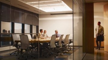 Three colleagues in a glass-walled meeting room, with a fourth person entering through the corridor