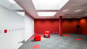 A modern office lounge with bold red walls, two red leather chairs on grey carpet, and a frosted glass partition