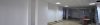 A mostly empty office space with white walls, tiled flooring, ceiling panels, and a stepladder near a lone desk in the background