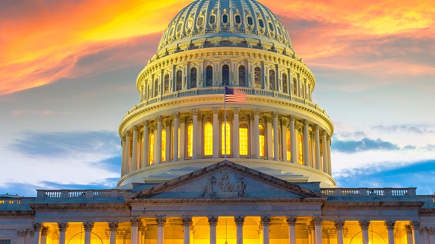 The illuminated US Capitol Building dome against an orange and blue sunset sky, with an American flag flying at its centre