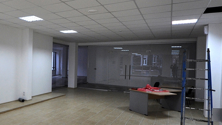 A mostly empty office space with white walls, tiled flooring, ceiling panels, and a stepladder near a lone desk in the background