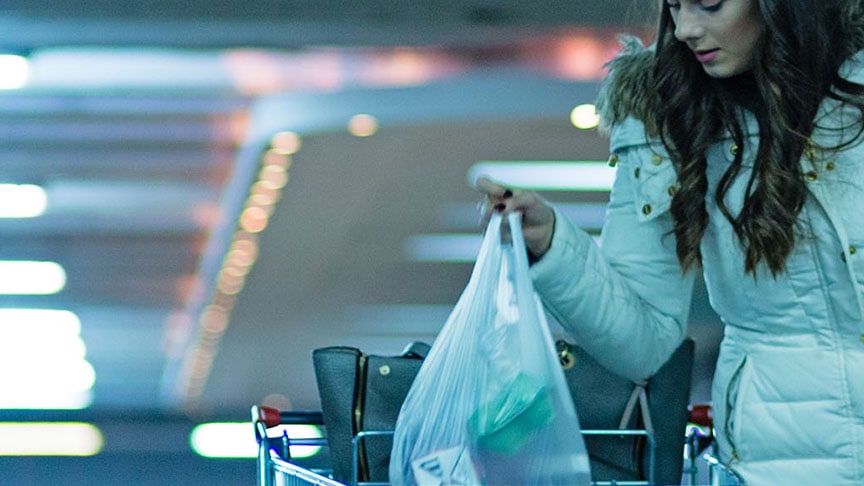 A person in a winter coat loading shopping bags from a trolley into a car boot in a car park