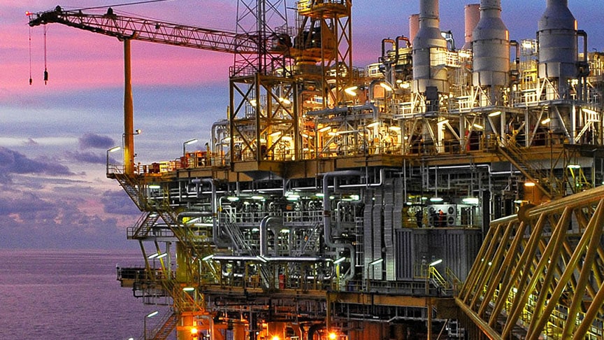 An illuminated offshore oil and gas platform with cranes and industrial structures, set against a vivid pink and purple sunset over the sea