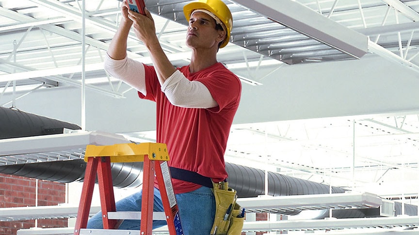 A worker with a yellow hard hat on a ladder, installing ceiling infrastructure in a large commercial building